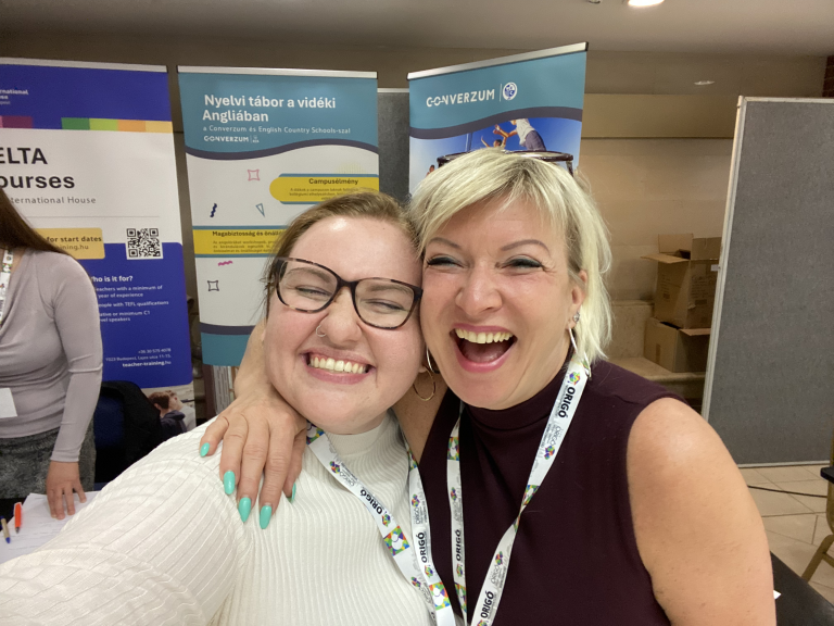 Two smiling women at a Budapest language education event, standing in front of course banners.