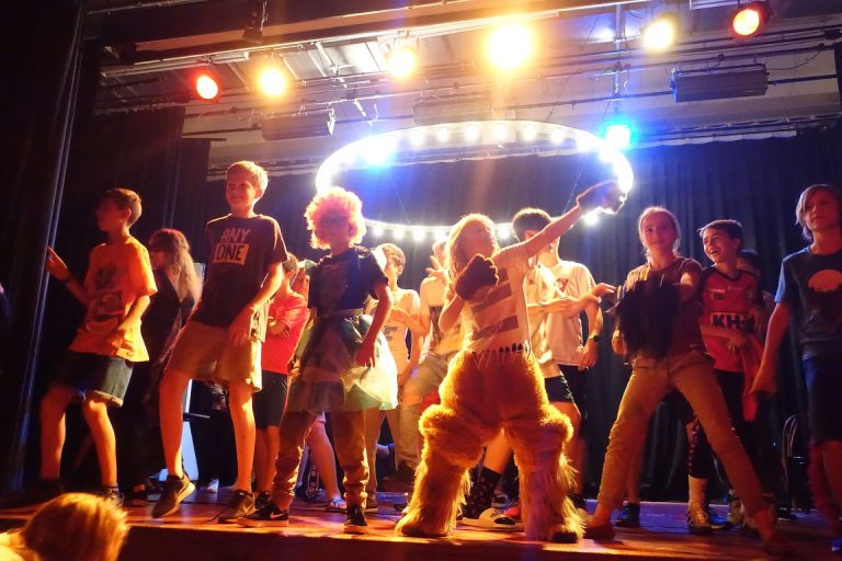 Children perform on stage under bright lights during an English summer camp talent show.